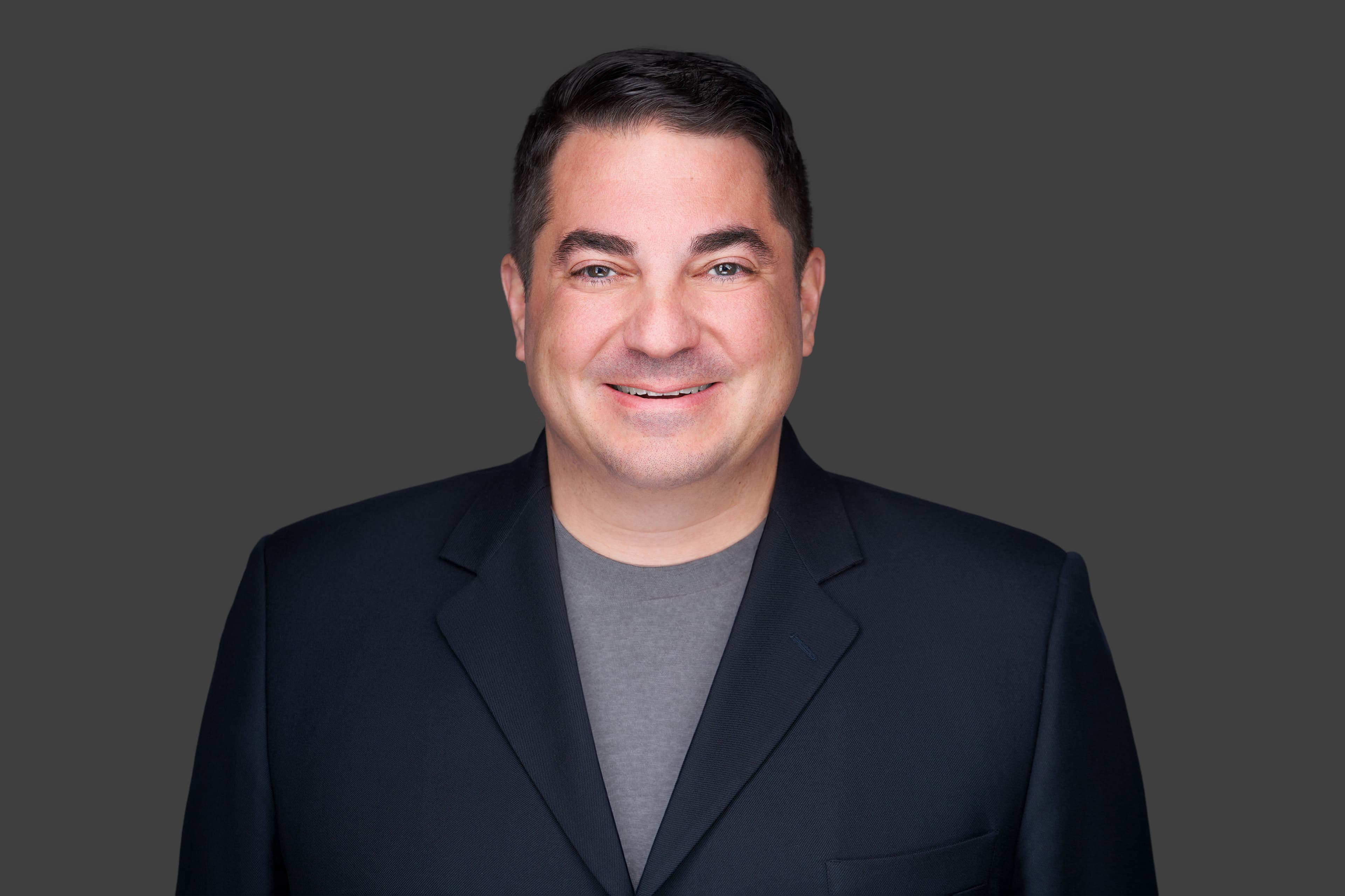 Portrait of a smiling man wearing a dark blazer over a dark shirt.