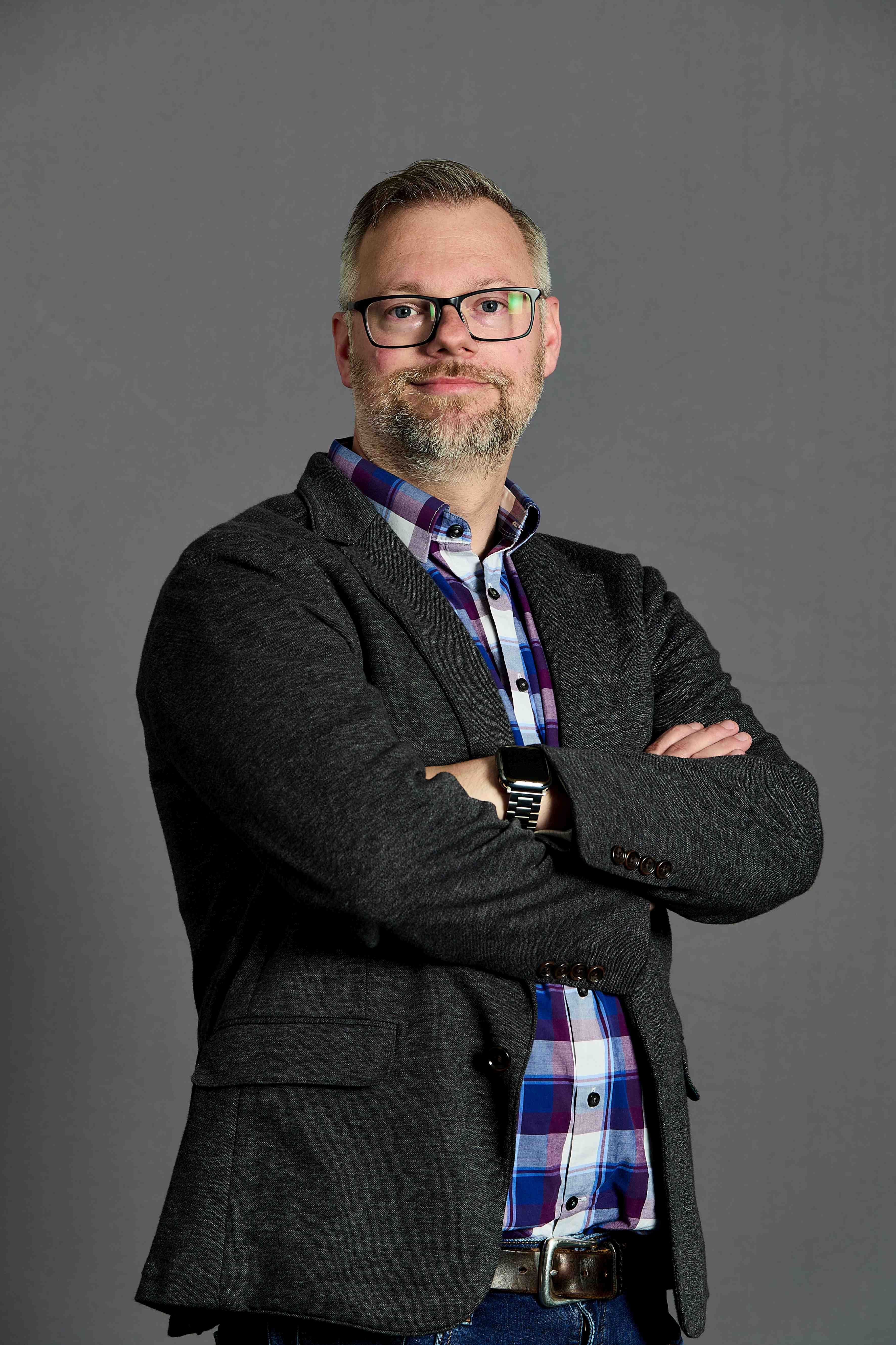 Portrait of a man wearing glasses, a dark jacket, and a plaid shirt with his arms crossed.