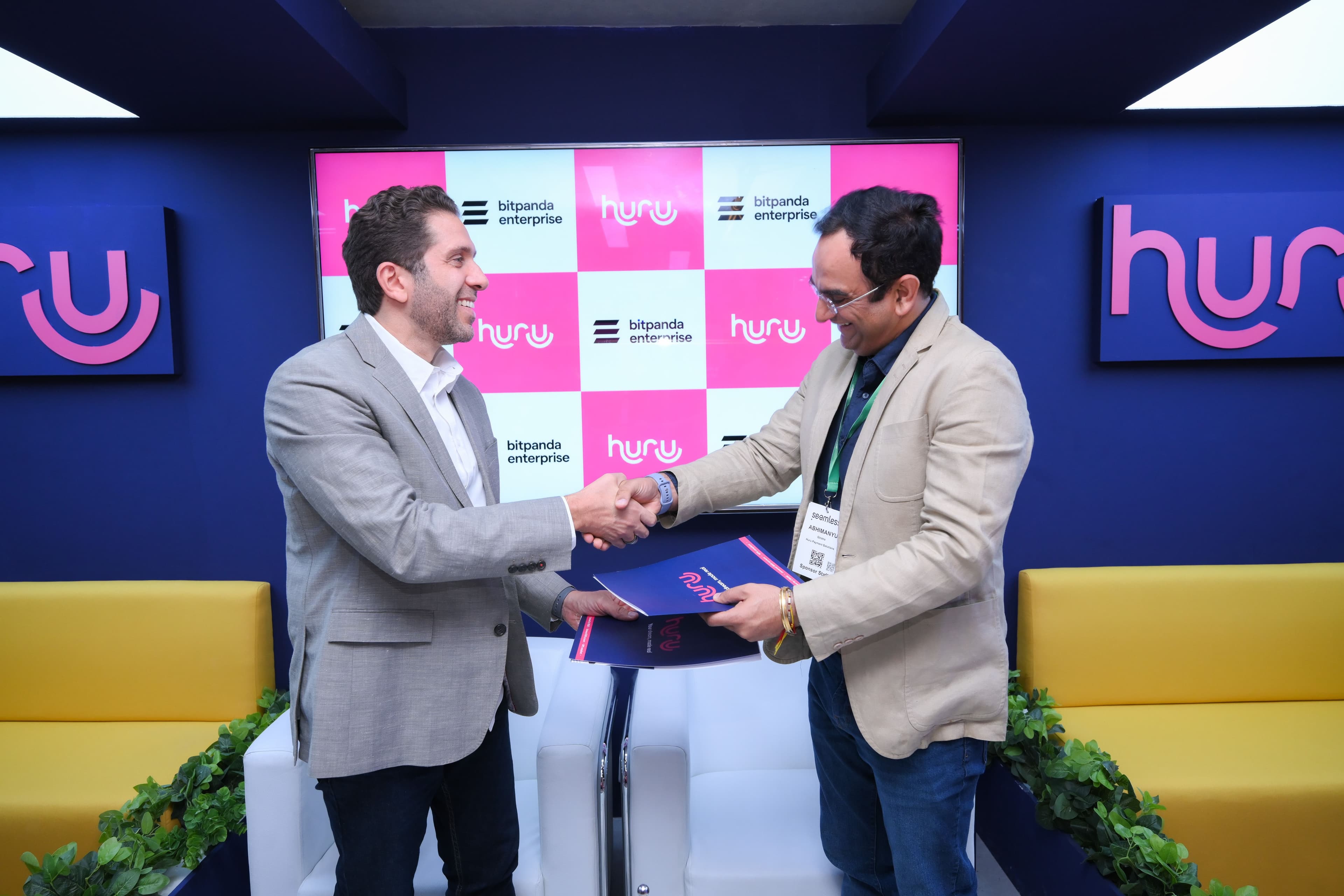 Two men shaking hands while holding agreement folders in front of Huru and Bitpanda Enterprise branding.