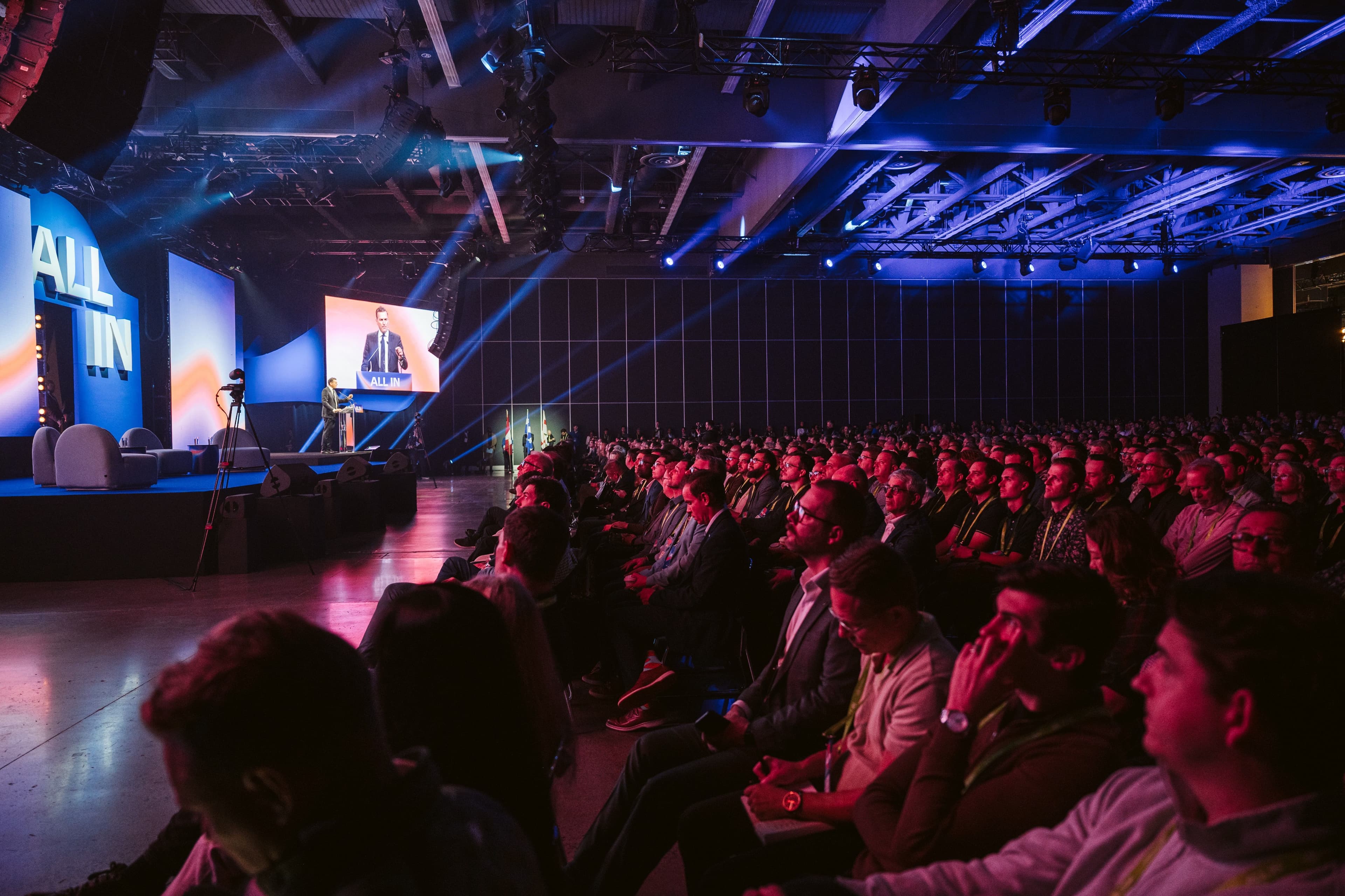 Audience seated in a dark auditorium facing an illuminated AI presentation stage.