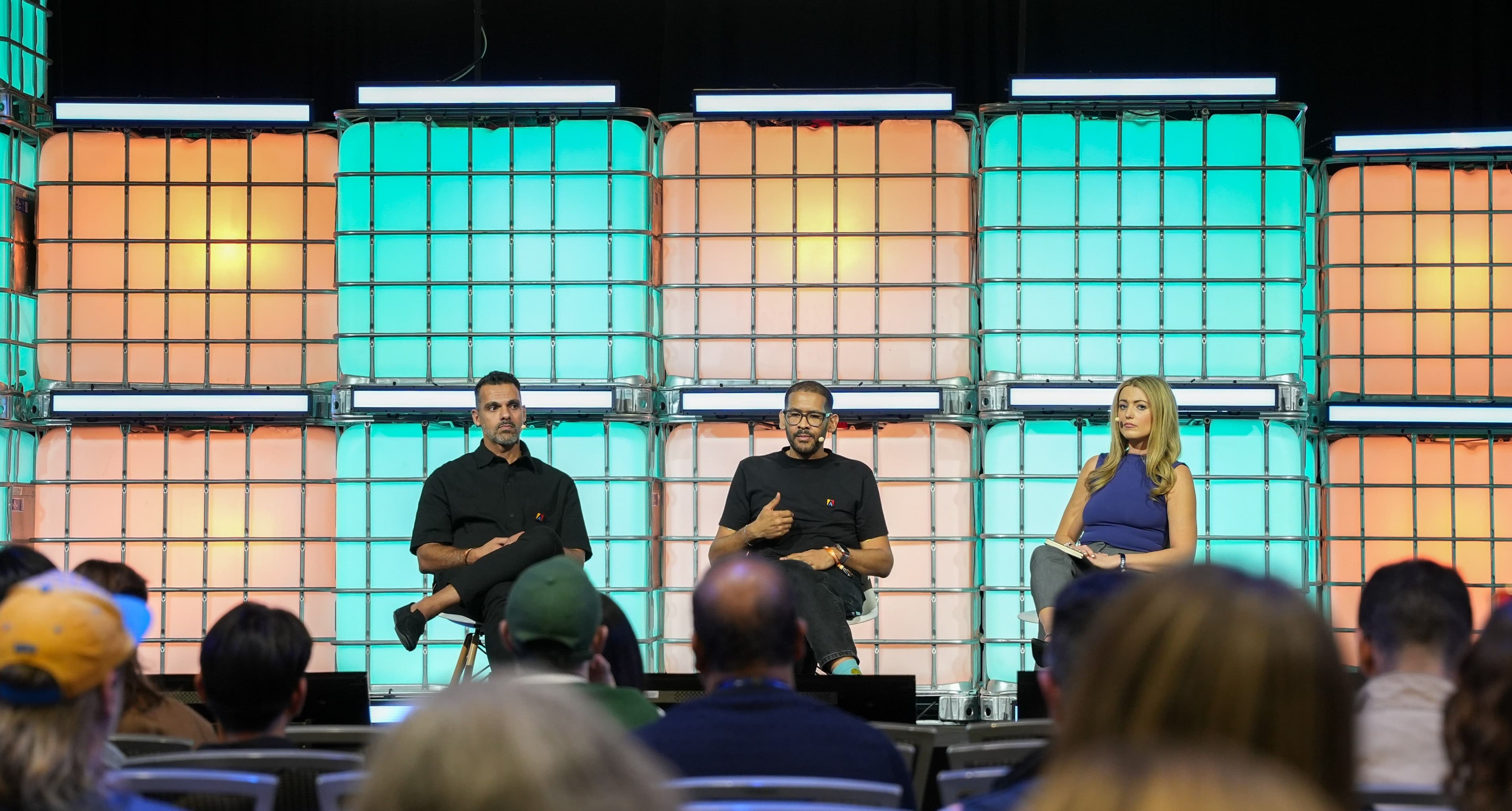 Three speakers seated on a stage in front of a colorful tiled display.