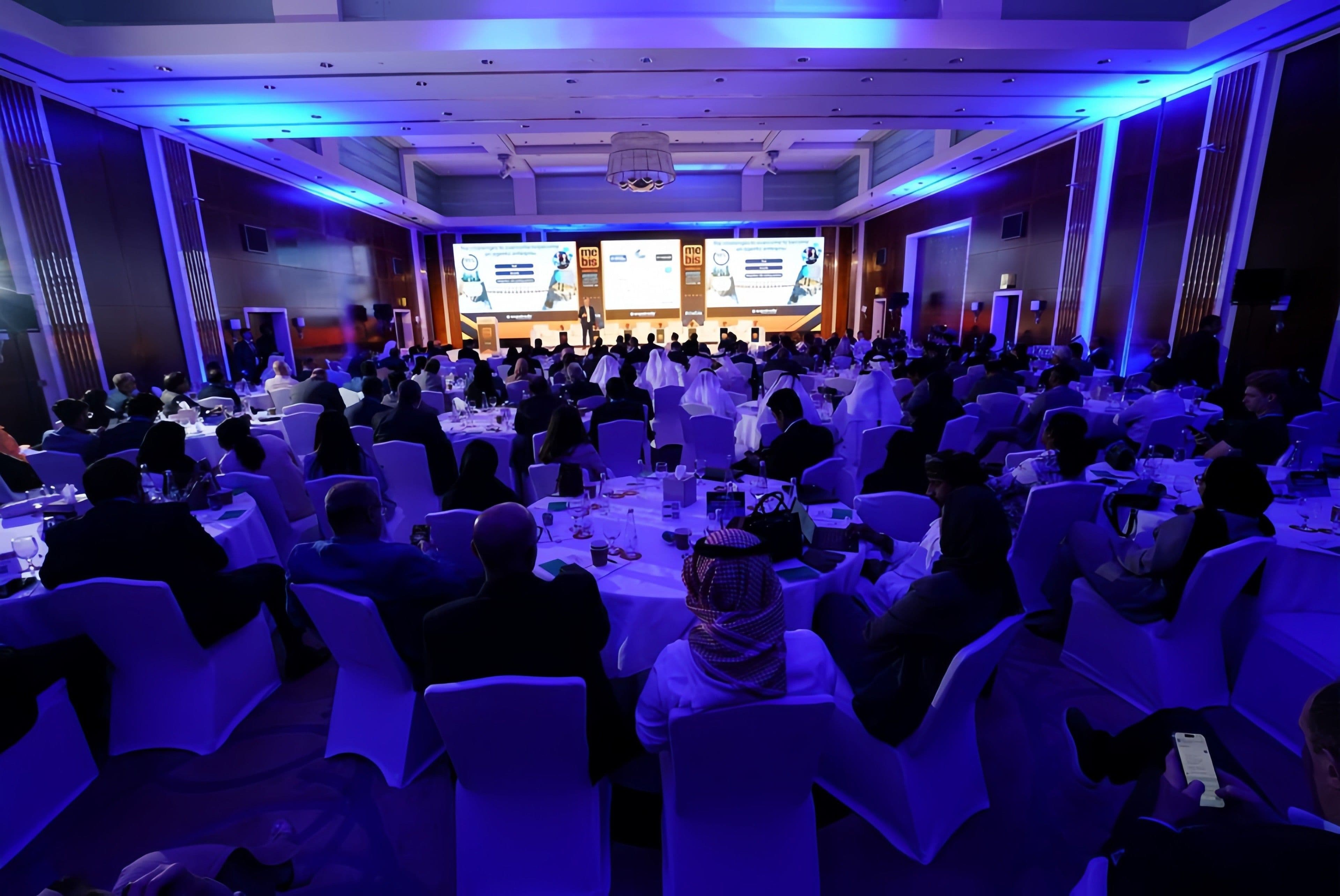 Audience seated in a blue-lit conference hall facing a stage.