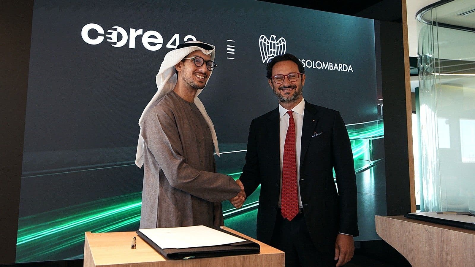 Two representatives shaking hands beside signed documents at an agreement ceremony.