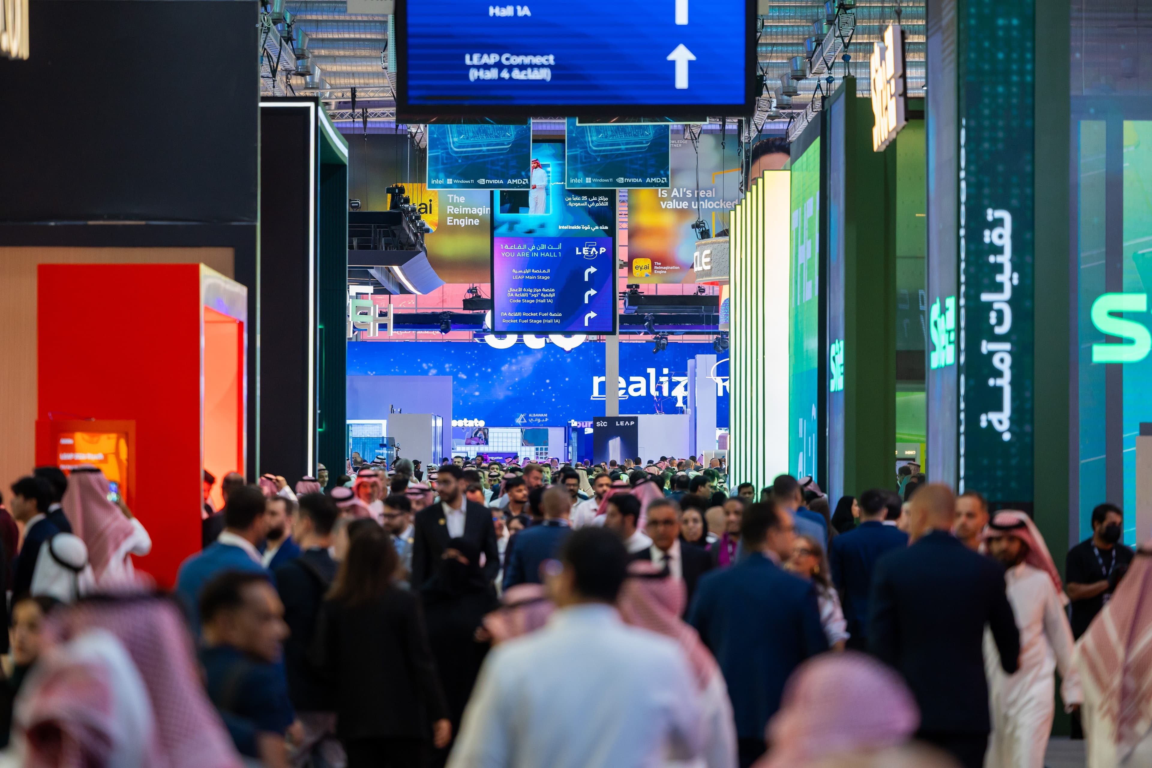 Visitors walking through a busy exhibition aisle lined with company signs.