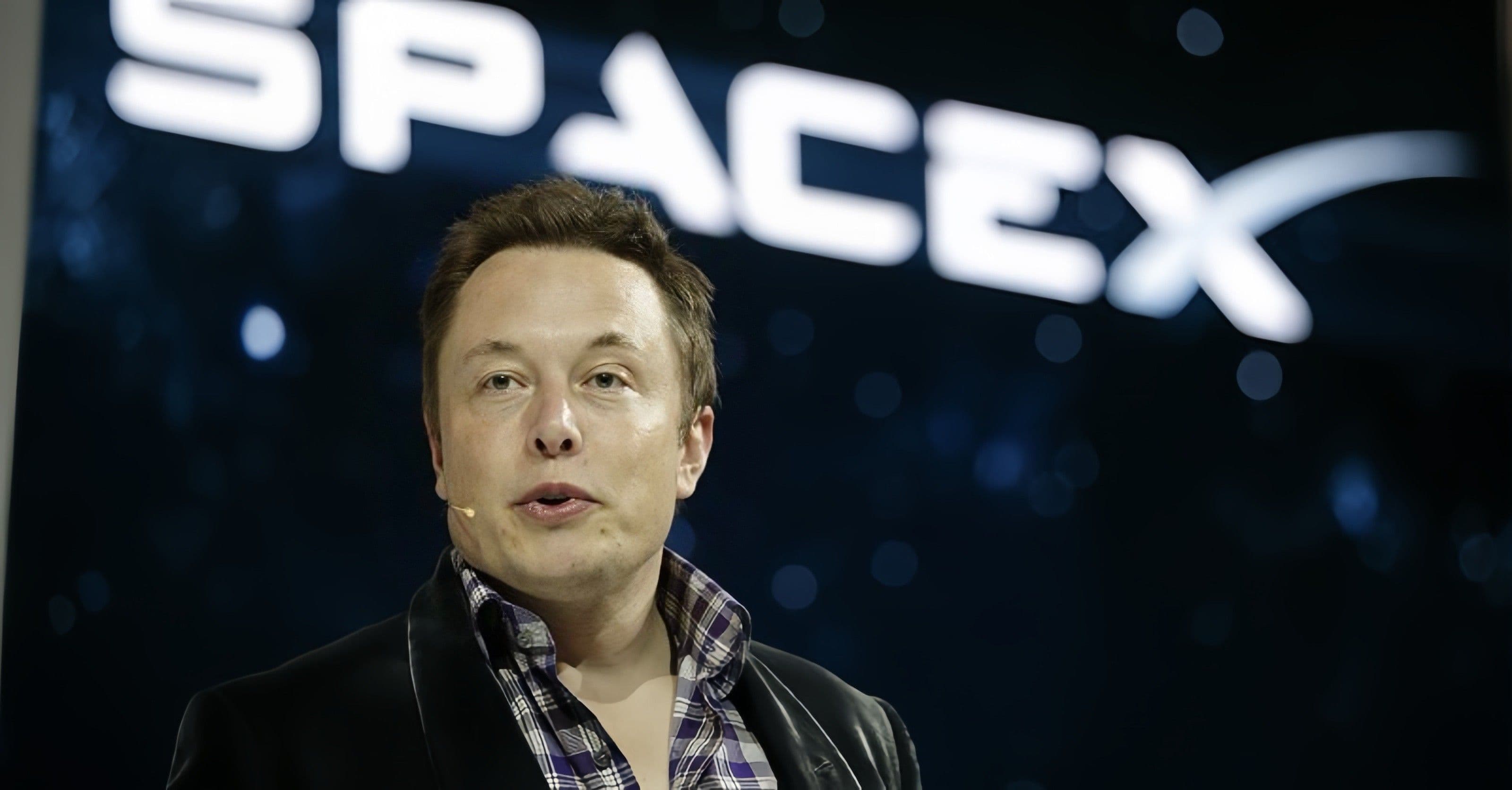 Elon Musk wearing a headset microphone in front of a SpaceX sign.